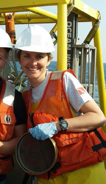A person in a hard hat and life jacket holds a sieve and stands in front of equipment on a boat. Another person is just out of frame.