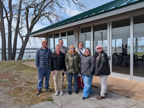 Seven people pose for a picture outside a small building with floor-to-ceiling windows at the waterfront.