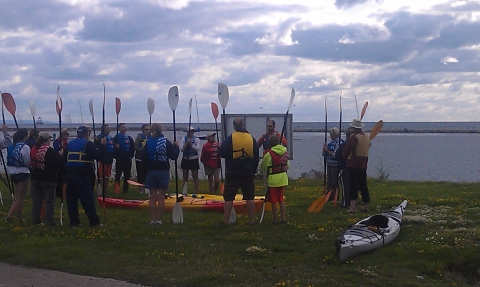 A group of people in life jackets stand on the shore with paddles near some kayaks.