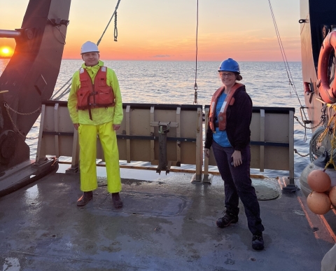 Two people wearing hard hats and life jackets stand on the back deck of a large boat on a lake at sunrise