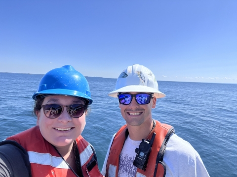 Two people wearing hard hats, life jackets, and sunglasses pose for a selfie in front of a large body of water on a cloudless sunny day