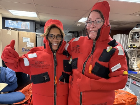 Two people try on emergency floatation suits while inside a lab on a boat