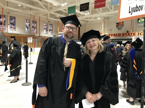 Two people dressed in academic regalia, including black gowns, black caps, and one of the people has a colorful hood draped over their arm. The other person's cap and gown are fancier, indicating a higher degree. In the background are other people in caps and gowns in a gymnasium.