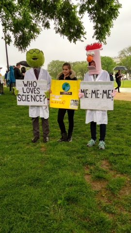 Three people holding signs. Two have lab coats and masks on resembling a green muppet with glasses (Bunsen) and a tall-headed muppet with bright red hair (Beaker). Bunsen holds a sign that says "Who loves science?" Beaker's sign says "Me-me-me!" A person stands between them with another sign.
