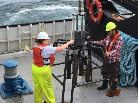 Two people stand on the deck of a large boat next to a weighted frame that has four tubes full of sediment.