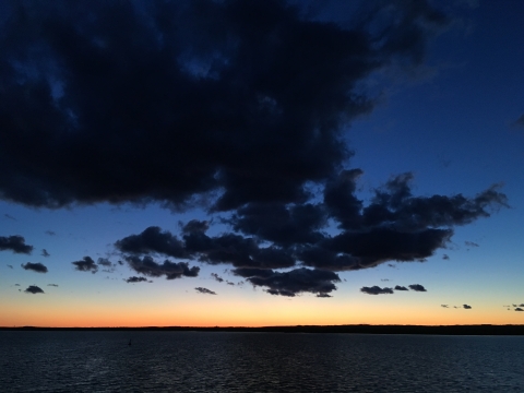 A dramatic picture of black clouds in a darkening sky. The sky fades from dark blue to a narrow band of yellow and orange right at the horizon, over the water.