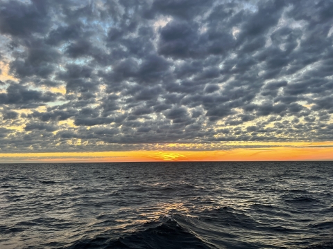 Sunset on a large body of water. A thin band of orange rests between gray dappled clouds and steely waves.