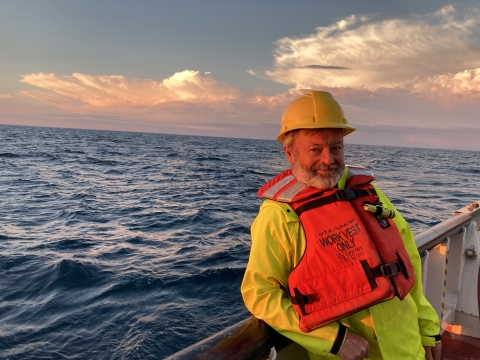 A person wearing a hard hat, life jacket, and rain gear poses near the railing of a boat in a large body of water with luminous clouds behind him.