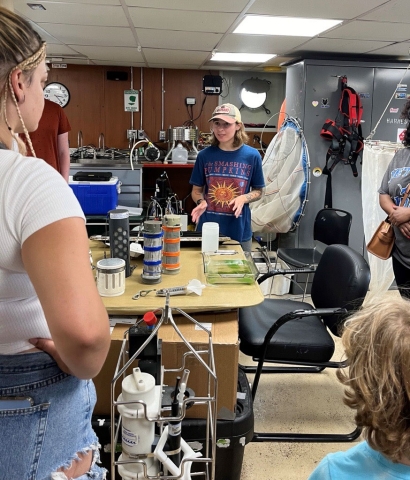 A person in a baseball hat stands by a table with scientific sampling equipment talking to visitors to a lab on a boat