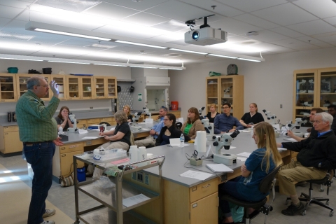 A person stands in front of a laboratory classroom giving a presentation. A group of people sit at the lab benches with microscopes.