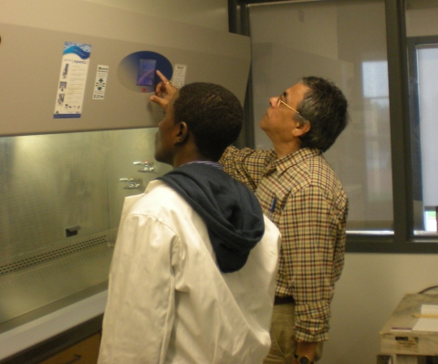 Two people standing in front of a fume hood. One is pressing buttons on the control panel above the fume hood.