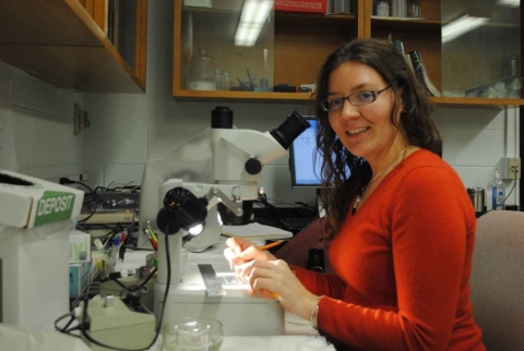 A light-skinned person with glasses smiles at the camera while working at a dissecting microscope in a lab