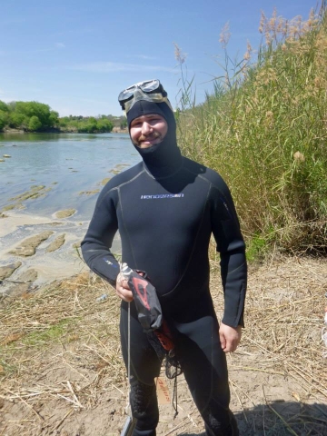 A person in a wet suit with goggles on their forehead stands by the bank of a river. Tall reeds are growing on the bank behind them.