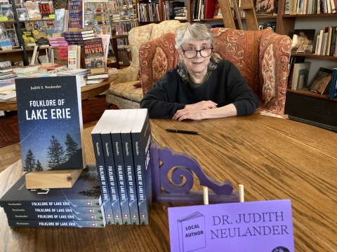 A person sits in an armchair behind a table with a stack of books "Folklore of Lake Erie" in a bookstore with a pen beside them for signing; tag "Local author Dr. Judith Neulander"