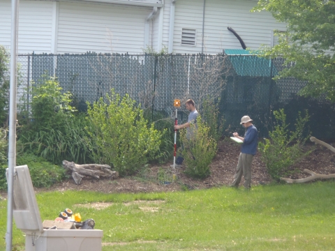 Two people working by a fence. One person holds surveying equipment while the other writes something down in a notebook.
