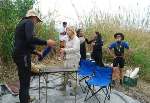 Several people set up a work station in a field with a portable table and chairs and field sampling equipment. Several of the people wear wetsuits.