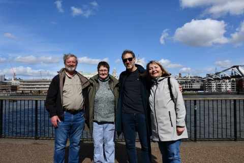 Four people pose for a picture by a river. They are wearing light jackets and the sky is bright with light clouds