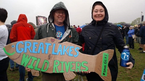 Two people in rain coats pose for a picture amidst a crowd of people. One is holding a sign shaped like a fish that says "I [heart] Great Lakes & Wild River #SaveGLRI"