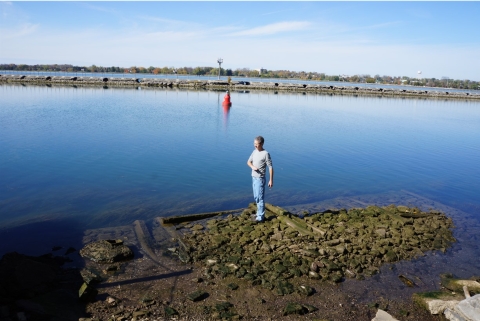 A person stands on algae-covered stone and wood cribbing by the water, gesturing  at waist level.