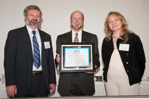 Three people pose for a picture. The person in the center holds up a framed certificate.