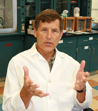 A person with short hair wearing a lab coat gestures with their hands while talking in a laboratory