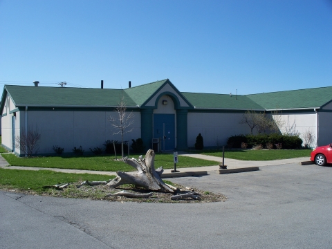 a small one story building with two wings, a central door, no windows, and an adjacent parking lot