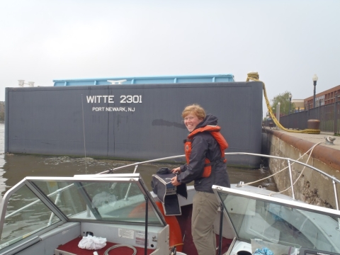 A person standing at the front of a boat tied to a wall. In front of the boat is a large freight boat marked "WITTE 2301 Port Newark, NJ"