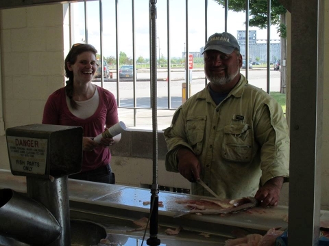 Two people stand inside a building at a counter where one of them is cutting up a fish.
