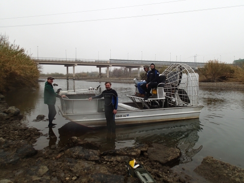Two people on an air boat while two people stand by it in very shallow water. A bridge crosses the river in the distance.