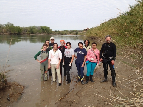 Ten people stand in shallow water near a river.