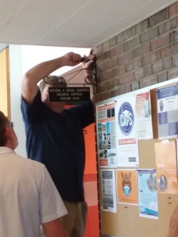 A person watches as another person uses a screwdriver on a sign on the wall of a hallway near a bulletin board. The sign says "Natural and Social Sciences Dean's Office Room A113."