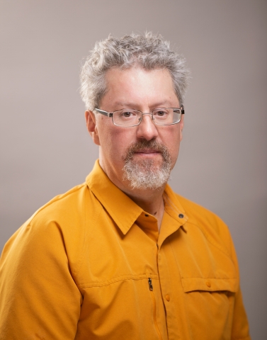 A ruddy light-skinned man with graying hair and beard wearing glasses and a yellow-orange buttoned shirt