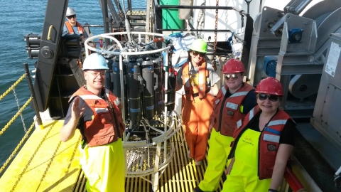 Four people in hard hats, rain pants, and life jackets stand on the deck of a boat near a large grey instrument.