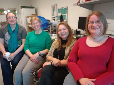Four people with long hair sit in chairs and smile in the lab.