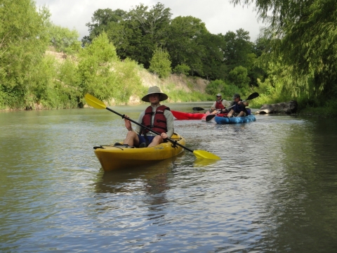 A person in a kayak in a narrow river. There are two other kayakers behind the first.