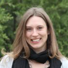 A smiling person with shoulder-length hair wearing a black scarf and a necklace while standing outside