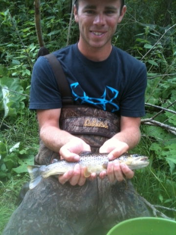 A person wearing chest waders holds a fish in their hands. They are standing in front of vegetation.
