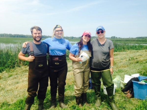 Four people wearing chest waders pose for a picture in a field near a wetland.
