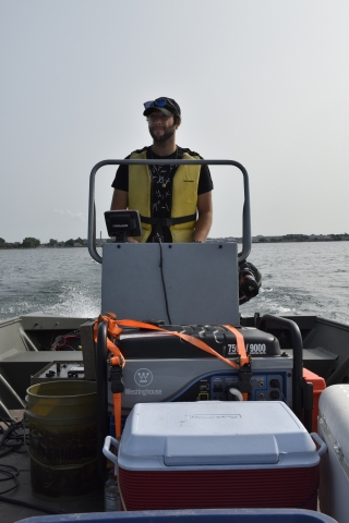 A person wearing a life jacket stands at the steering console of a small boat in a river.