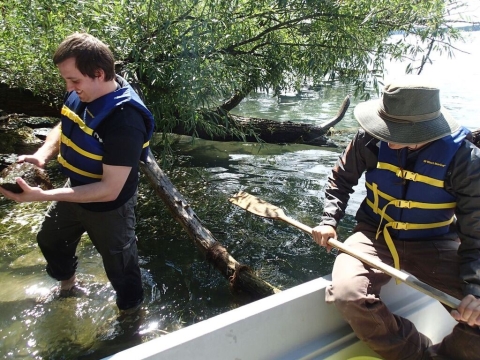 A person in a life jacket wads in shallow water holds a rock, next to a person on a small boat with an oar in their lap, face obscured by a hat.