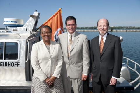 Three people in suits or a suit jacket over a dress stand in front of a boat at the waterfront.