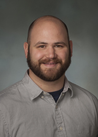 A balding person with a beard and moustache smiles, wearing a gray button-up shirt over a black t-shirt.