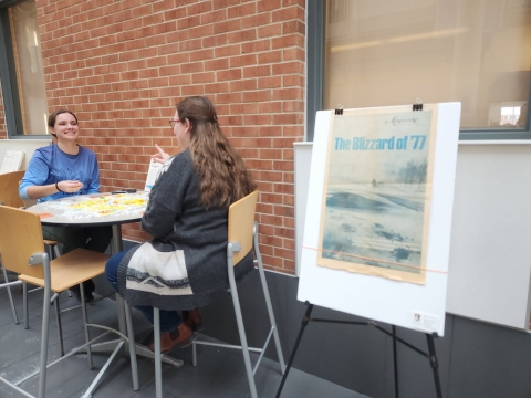 Two people sit at a cafe table playing a board game. A poster with a full page spread from a newspaper has a picture of snow and title "The Blizzard of '77" displayed on an easel
