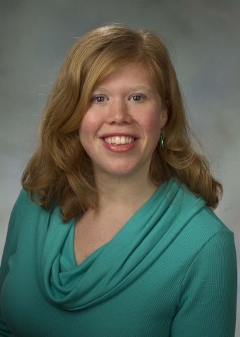 A person with long, wavy red hair in a side part smiles, wearing some makeup, a dangly green earring, and a teal cowl-neck blouse