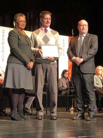 Three people dressed professionally stand together on a stage, the center one holding a plaque. Behind are banners and people seated in chairs.