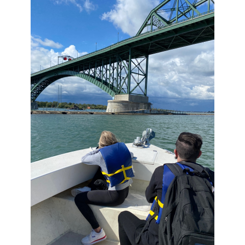 Two people sitting in the front of the boat, looking forward. The boat is in a canal next to a river and is about to go under a tall bridge.