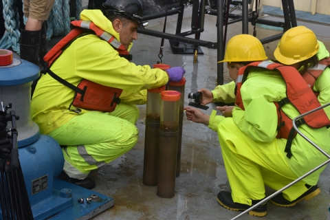 Three people in rain gear and safety gear crouch down on the deck of a ship around four tubes of mud and water and take pictures of them