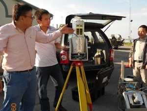 Three people stand around an instrument mounted on a tripod in a parking lot near a van