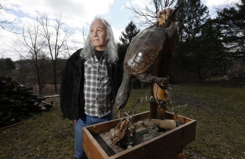a person with long gray hair, jeans, and an open jacket over a flannel shirt stands next to a diving taxidermied turtle on a natural stand while outdoors in a yard with evergreens and leafless deciduous trees