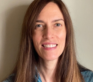 A smiling light-skinned woman with long brown hair waring a patterend blue shirt